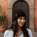 A woman wearing a black knit beanie with a small white rectangular logo patch, standing in front of a warm-toned stone archway and terracotta wall.