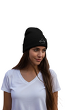 A woman wearing a black knit beanie with a small white embroidered Mexico City logo on the front, paired with a white V-neck t-shirt, set against a plain white background.