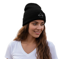 A woman with long wavy brown hair wearing a black knit Mexico City beanie with a small white logo on the front, paired with a white V-neck t-shirt, set against a plain white background.