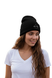 A woman with long wavy brown hair wearing a black knit Mexico City beanie with a small white logo on the front, paired with a white V-neck t-shirt, set against a plain white background.