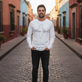 A man stands on a cobblestone street wearing a white long-sleeve crewneck sweatshirt with a black outline map graphic and the text 'Norte deñ' across the chest, paired with black jeans and black boots.