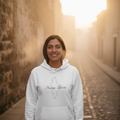A woman stands on a sunlit cobblestone street wearing a white hoodie with a black outline map graphic and the words 'Marea Loca' across the chest, paired with black pants.