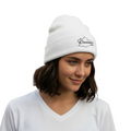 A woman wearing a white Oaxaca knit beanie with a black embroidered logo and a white V-neck t-shirt, standing outdoors in front of a warm-toned stucco building with a cactus in the background.