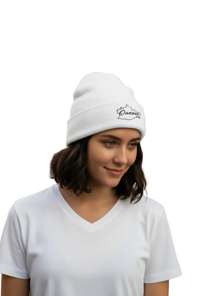 A woman wearing a white Oaxaca knit beanie with a black embroidered logo and a white V-neck t-shirt, standing outdoors in front of a warm-toned stucco building with a cactus in the background.