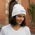 A woman wearing a white knit beanie with a black embroidered logo and a white V-neck t-shirt, standing outdoors in front of a warm-toned stucco building with a cactus in the background.