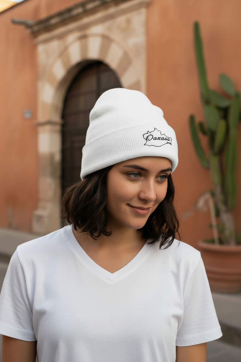 A woman wearing a white knit beanie with a black embroidered logo and a white V-neck t-shirt, standing outdoors in front of a warm-toned stucco building with a cactus in the background.