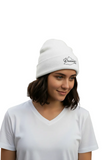 A woman wearing a white Oaxaca knit beanie with a black embroidered logo and a white V-neck t-shirt, standing outdoors in front of a warm-toned stucco building with a cactus in the background.