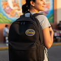 A woman wearing a gray t-shirt and blue jeans is shown from the back, carrying a black backpack with a circular 'Oaxaca' logo patch on the front. The background features a colorful mural on a brick wall, lit by warm golden hour sunlight.
