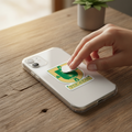 Hand pressing a die-cut bubble-free sticker of the official coat of arms of Yucatán, Mexico onto a white phone case — featuring a green and yellow shield with a deer and tree design symbolizing the state’s heritage and nature.