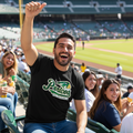 A man in a baseball stadium wearing a black t-shirt with a green and white 'Pericos' logo, cheering with a raised fist. Other spectators are seated around him in the stands, with the baseball field visible in the background.