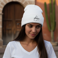 A woman wearing a white knit beanie with a black embroidered logo that reads 'La Piel de Miel' poses outdoors in front of a warm-toned stone building and cactus.