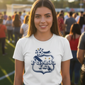 A smiling young woman stands on an outdoor sports field at sunset wearing a white short-sleeve T-shirt with a navy blue 'Puebla FC' soccer crest graphic on the front, paired with blue jeans.