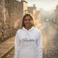 A smiling woman stands on a cobblestone street at sunset wearing a white hoodie with a black outline map of Puebla and the word 'Puebla' across the chest, paired with black pants.