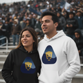 A man and a woman standing together in stadium seating, both wearing matching hoodies with a blue and gold graphic logo. The woman's hoodie is black, and the man's hoodie is white.
