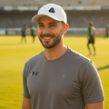 A smiling man on a sports field wearing a white Pumas UNAM cap and a gray Under Armour t-shirt, with stadium lights and seating in the background.