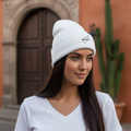 A woman wearing a white Queretaro ribbed knit beanie with a small black embroidered logo, standing outdoors in front of a terracotta-colored building and cactus.