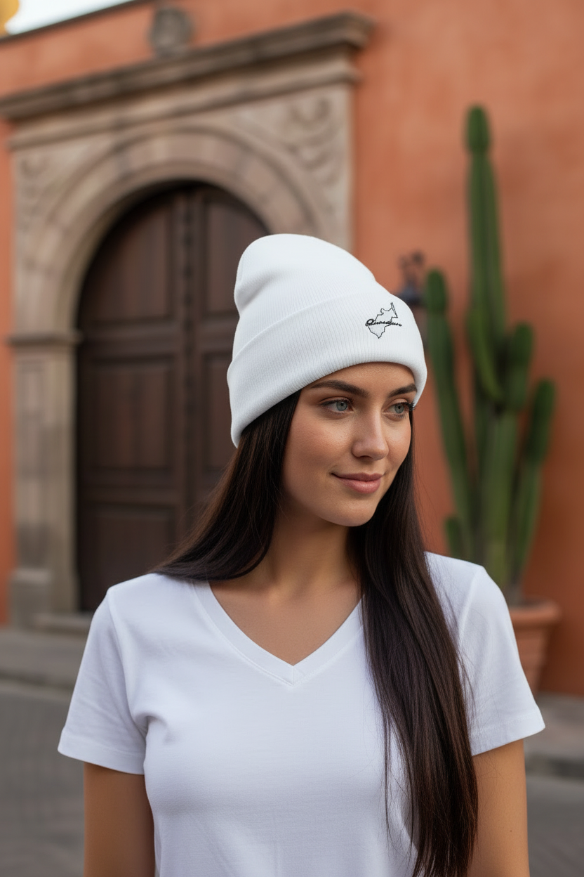 A woman wearing a white Queretaro ribbed knit beanie with a small black embroidered logo, standing outdoors in front of a terracotta-colored building and cactus.