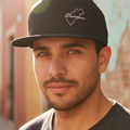 A man wearing a black baseball cap with a white outlined map graphic and the word 'Queretaro' on the front, photographed outdoors with warm sunlight and a blurred building background.