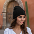 A woman wearing a Quintana Roo black knit beanie with a small white embroidered logo, standing outdoors in front of a warm-toned stucco building with a stone archway.