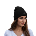A woman with long brown hair wearing a black knit beanie with a small white embroidered Quintana Roo logo, paired with a white V-neck t-shirt, set against a solid white background.