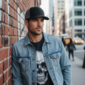 A man wearing a black baseball cap, a light blue denim jacket, and a black t-shirt with a large white skull graphic, standing against a red brick wall in an urban street setting.