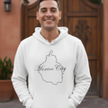 A smiling man wearing a white pullover hoodie with a black outline graphic of Mexico City and the text 'Mexico City' across the chest, standing in front of a rustic wooden gate and terracotta wall.
