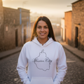 A smiling woman stands on a cobblestone street wearing a white hoodie with a black outline map of Mexico City and the words 'Mexico City' printed on the front. The background shows a softly blurred outdoor town scene at sunset.