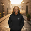 A woman stands in a sunlit cobblestone alley wearing a black hoodie with a small white script logo on the left chest. The background is softly blurred with warm golden light.