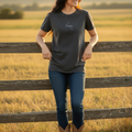 A woman stands outdoors in a field behind a wooden fence, wearing a dark charcoal heather t-shirt with a small white script logo on the chest, blue jeans, and brown cowboy boots.