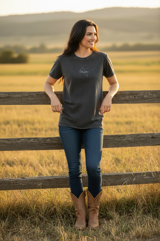 A woman stands outdoors in a field behind a wooden fence, wearing a dark charcoal heather t-shirt with a small white script logo on the chest, blue jeans, and brown cowboy boots.
