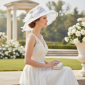 A woman in a white lace wedding dress and large white hat sits outdoors in a garden setting, holding a small white clutch.