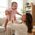 A woman in a cream long-sleeve top and blue jeans holds a smiling baby up in a bright living room. The baby is wearing a light pink onesie with the text 'Just One More' in dark script.