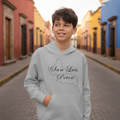 A smiling young boy stands on a cobblestone street wearing a light gray hoodie with a black outline map of Colombia and the text 'San Luis Polot' printed on the front. The background shows colorful colonial-style buildings in a street scene.