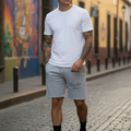 A man standing on a cobblestone street wearing a white crew-neck t-shirt and light gray sweat shorts with 'You Can Fly!' printed on the left leg.