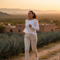 A woman jogging outdoors on a dirt path at sunset, wearing a white long-sleeve athletic top and light beige jogger pants with white sneakers. Agave plants and tan buildings are visible in the background.