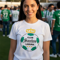 A smiling young woman stands on a sports field wearing a white Club Santos Laguna t-shirt with a green and gold soccer ball graphic and crown logo.