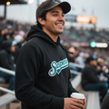A man at a stadium wearing a black hoodie with a teal and white 'Saraperos' logo on the chest, a dark baseball cap, and holding a coffee cup.
