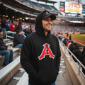 A man wearing a black Acereros de Monclova hoodie with a large red and white 'A' logo on the front stands in a stadium crowd, smiling. The background shows a baseball field and stadium seating.
