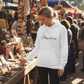 A woman wearing a white hoodie with 'Sinaloa' and a map outline printed on the front, standing at a rustic market stall in a sunny outdoor market.