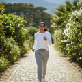 A woman jogging on a cobblestone path surrounded by lush green bushes and distant mountains, wearing a white long-sleeve top, light gray joggers, and white sneakers.
