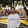 A man wearing a white long-sleeve t-shirt with a black outline of the state of Mexico and the word 'Sinaloa' written across it, standing outdoors in a park-like setting with trees and a stone building in the background.