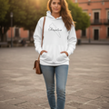 A woman stands in a sunlit outdoor plaza wearing a white pullover hoodie with a black outline graphic of the island of Jamaica and the word 'Jamaica' printed across the chest. She is styled with light blue skinny jeans, brown ankle boots, and a brown shoulder bag.