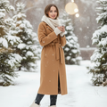 A woman stands in a snowy outdoor setting wearing a camel-brown belted coat, a cream scarf, dark leggings, and cream ankle boots. Snow-covered evergreen trees and soft hanging lights are visible in the background.
