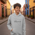 A smiling young boy stands on a cobblestone street wearing a light gray hoodie with a black outline of the state of Mexico and the word 'Sonora' printed across the chest. Colorful colonial-style buildings line both sides of the street in the background.