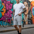 A man wearing a white crewneck t-shirt and light gray drawstring shorts with 'Chambriard' printed on the left leg, standing on a cobblestone street in front of a colorful graffiti wall.