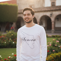 A smiling man wearing a white long-sleeve t-shirt with a black outline map of Sonora and the word 'Sonora' written in script, standing outdoors in front of a historic building with arches and flowers.