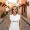A woman stands on a cobblestone street wearing a white sleeveless tank top with a black outline of the state of Sonora and the word 'Sonora' printed across the front. The background shows a narrow street lined with warm-toned buildings and hanging flower baskets.
