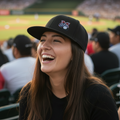 A woman smiling in a stadium crowd, wearing a Sultanes de Monterrey black baseball cap with a small multicolor logo and a black t-shirt. The background shows a blurred baseball field and stands.