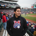A man wearing a black pullover hoodie with a 'Sultanes' graphic on the chest stands in a stadium crowd during a baseball game.
