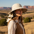 A woman wearing a wide-brimmed beige sun hat with a neck flap and a brown vest over a white embroidered blouse, standing in a dry grassy landscape with red rock formations under a clear blue sky.